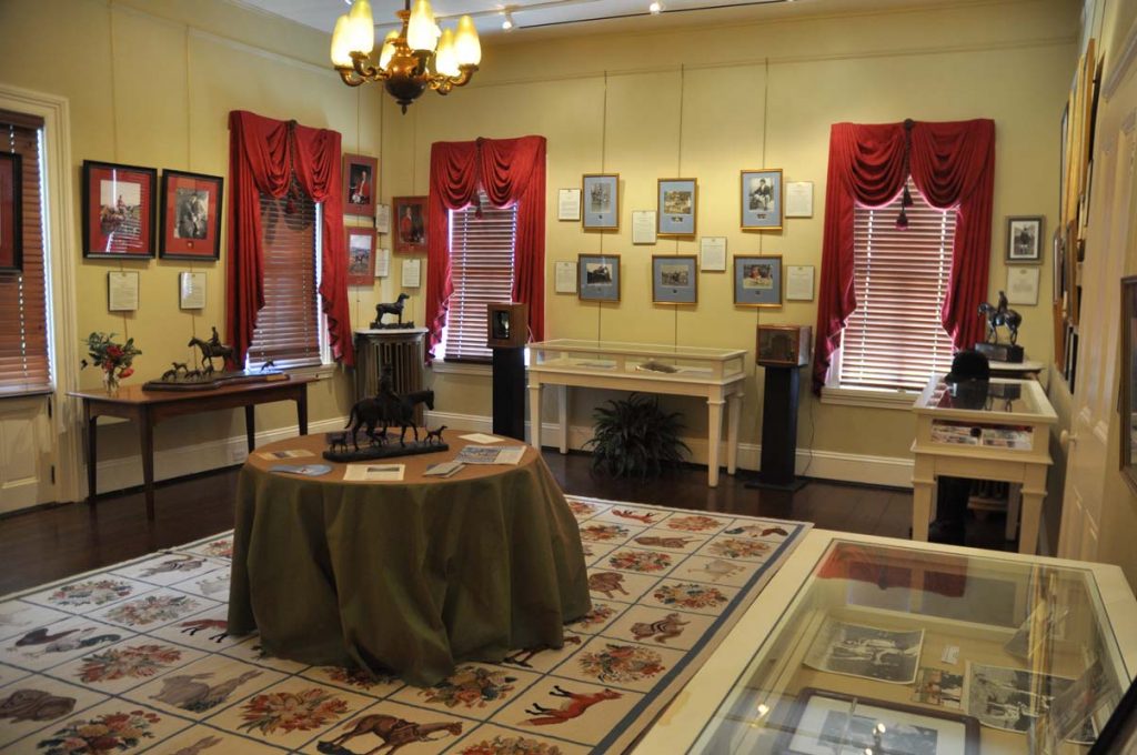 Exhibit room at the MHHNA with framed art on the walls, a fire place, and a table.
