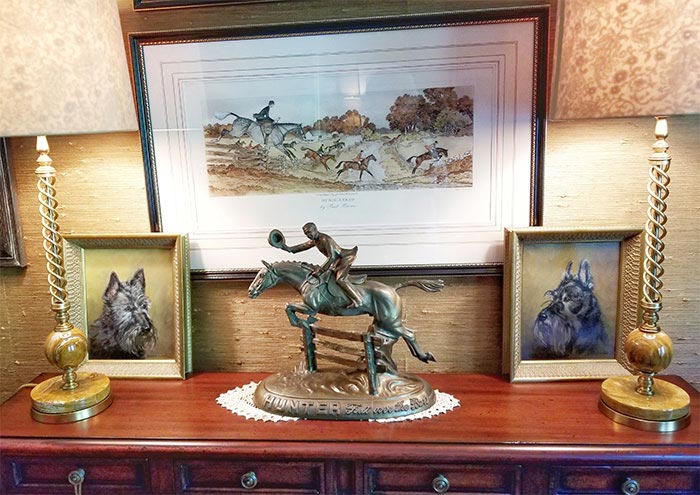 Table with two lamps, two painted portraits of dogs, a bronze foxhunting sculpture, and a framed illustration of a foxhunting scene on the wall.