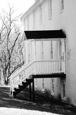 Old black and white photo of a three story white building with steps and a porch leading to the second floor.